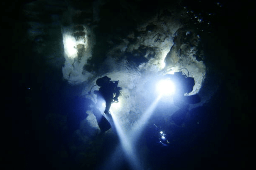 Two scuba divers explore a dark underwater cave, illuminating rocky walls with bright torches as they swim side by side, surrounded by deep blue water.