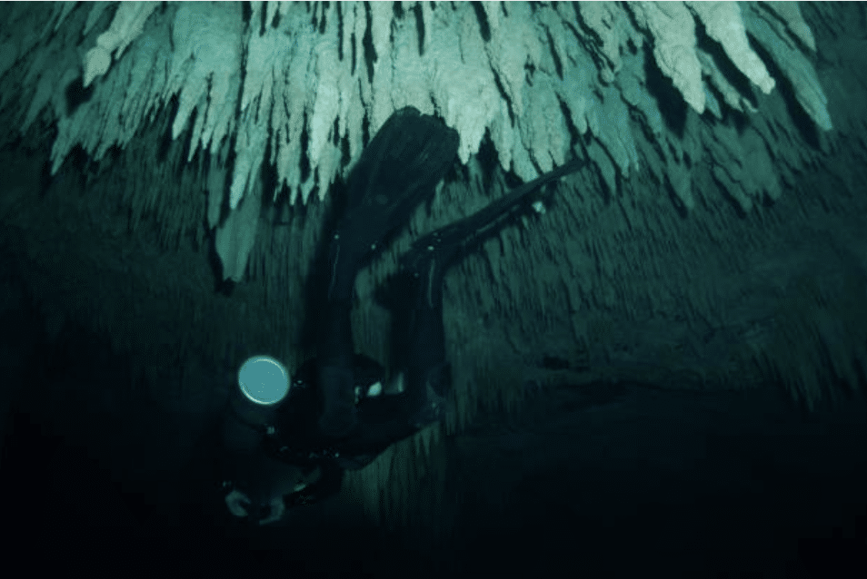 A scuba diver swims underwater near sharp, jagged stalactites hanging from the cave ceiling in a dark, submerged cavern.