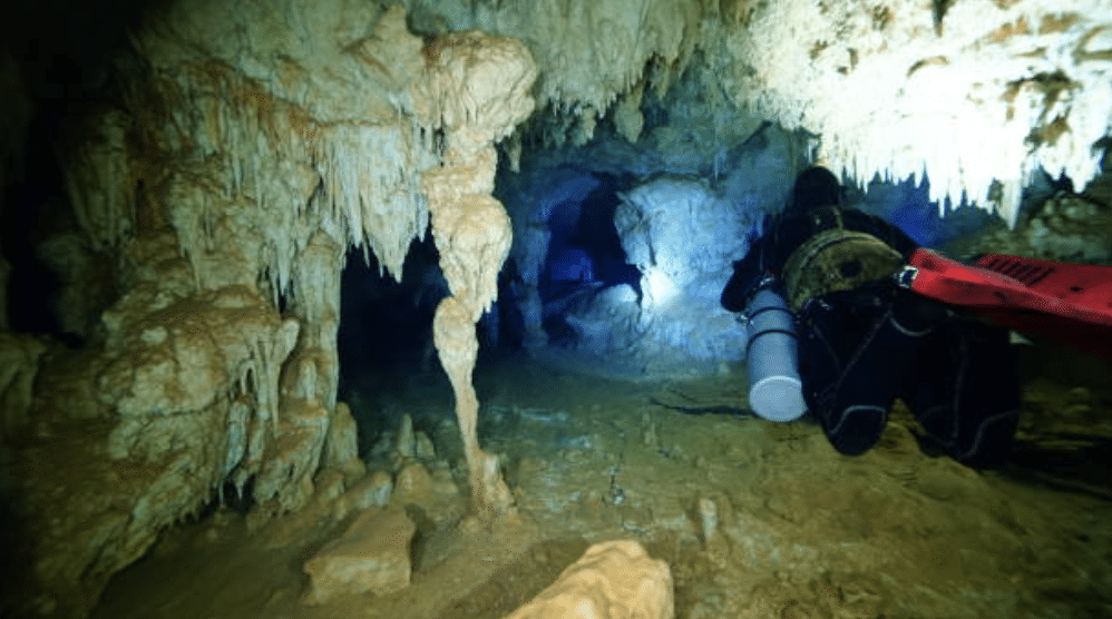Screenshot 2026-03-15 at 210312 - N9BO℠ | Global Underwater Services Ltd A scuba diver with a torch explores an underwater cave filled with stalactites and stalagmites, surrounded by rock formations and dim, blue-tinged water.