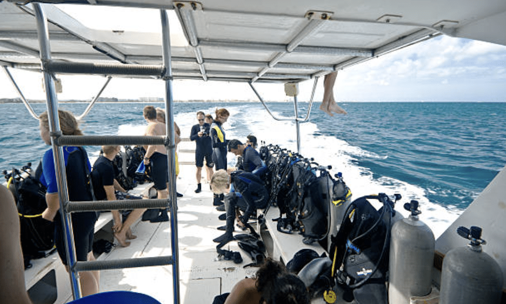 Screenshot 2026-03-15 at 215254 - N9BO℠ | Global Underwater Services Ltd A group of people in wetsuits prepare for scuba diving on a boat deck, surrounded by scuba tanks and gear, with the sea and sky visible in the background. One person sits above the deck with feet dangling.