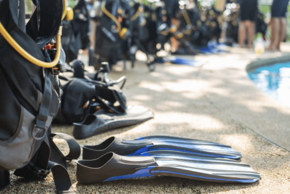Screenshot 2026-03-15 at 215312 - N9BO℠ | Global Underwater Services Ltd A row of scuba fins and diving gear is laid out on the ground beside a swimming pool, with groups of people standing in the background.