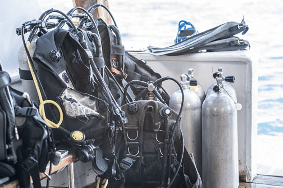 Screenshot 2026-03-15 at 215408 - N9BO℠ | Global Underwater Services Ltd Two scuba diving gear sets, including cylinders, regulators, and buoyancy control devices, rest on a wooden bench next to three spare cylinders and a pair of fins on a boat with water visible in the background.