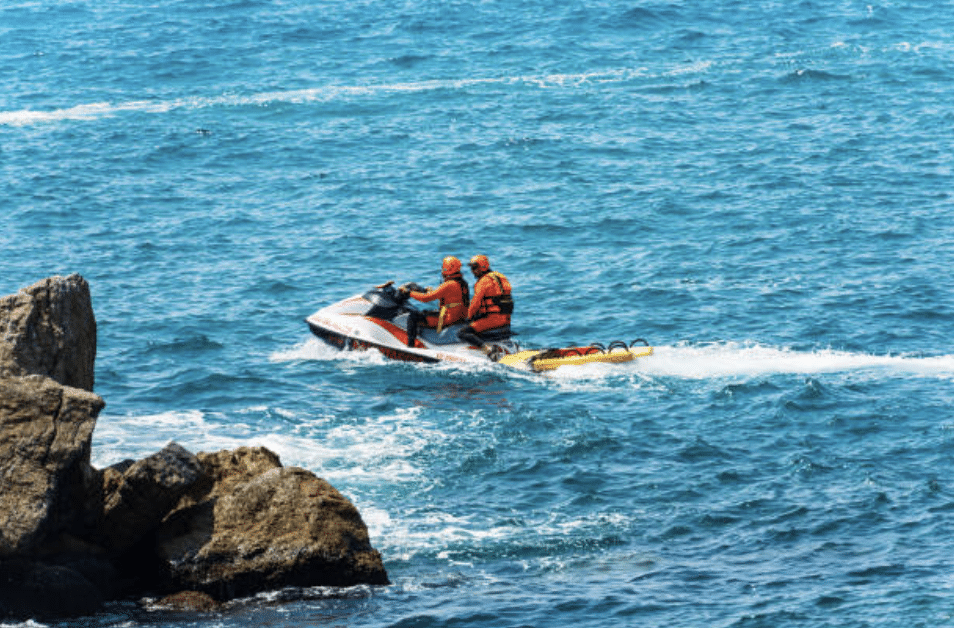 Two people wearing orange life jackets and helmets ride a red and white jet ski on bright blue water near some large rocks, with a yellow rescue board attached to the back.