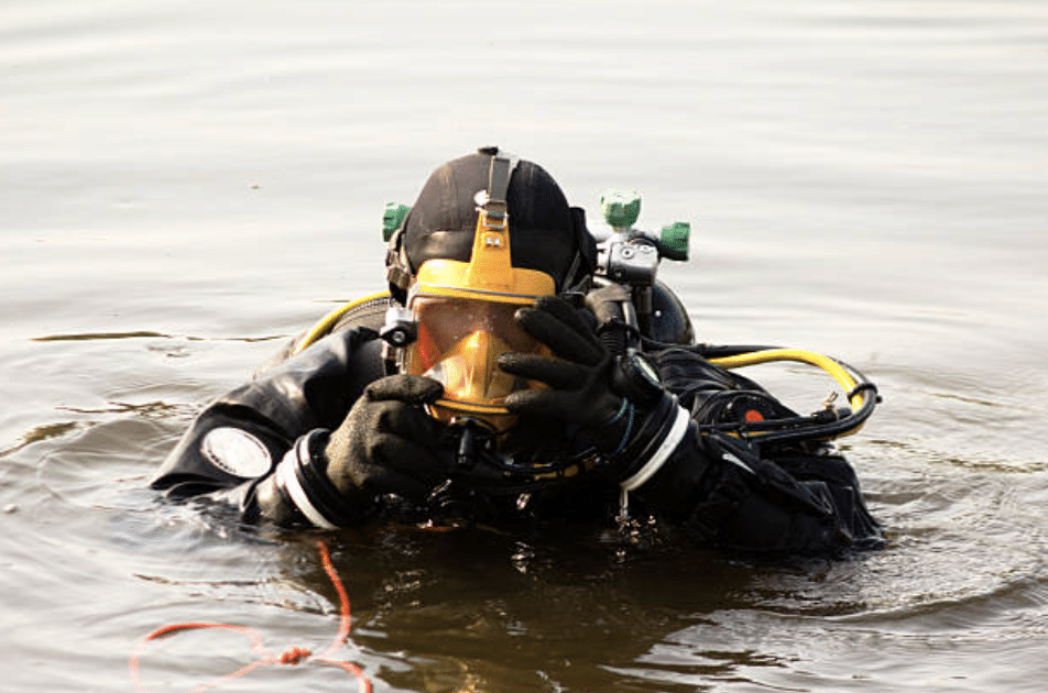 A diver wearing a full-face mask and black wetsuit is partially submerged in water, adjusting the mask with both hands. Scuba gear and hoses are visible on their back.