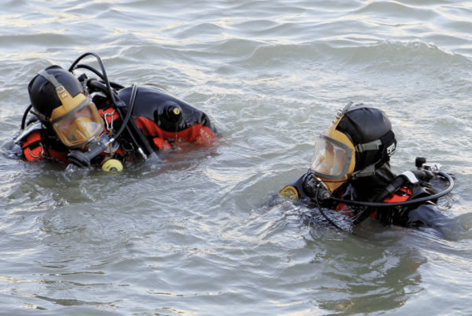 Two scuba divers wearing full-face masks and wetsuits float in choppy water, appearing to communicate or prepare for an underwater activity.