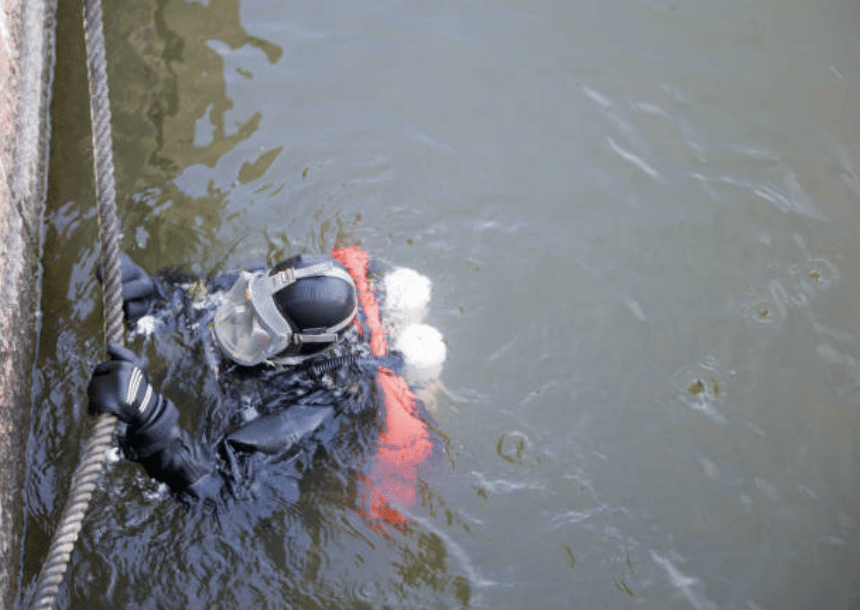 A diver wearing a black wetsuit, gloves, and mask holds onto a rope whilst partially submerged in murky water, with an orange vest and air bubbles visible around their head.