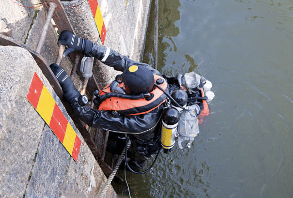 A diver in a wetsuit and scuba gear climbs a metal ladder out of the water onto a concrete quay with yellow and red warning stripes.