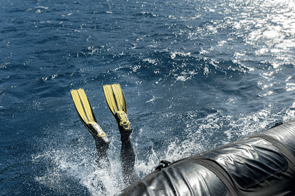 Screenshot 2026-03-15 at 225559 - N9BO℠ | Global Underwater Services Ltd A person wearing yellow flippers dives headfirst into the blue sea near an inflatable boat, with just their legs and flippers visible above the water.