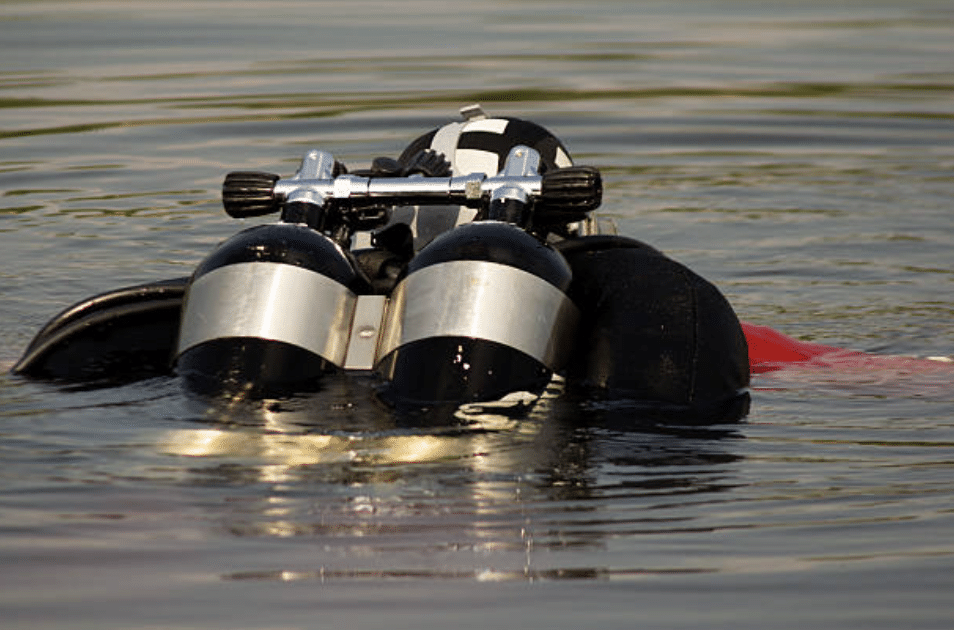 Screenshot 2026-03-15 at 225607 - N9BO℠ | Global Underwater Services Ltd A scuba diver wearing a black wetsuit and twin air cylinders partially submerged in calm water, with equipment visible above the surface.