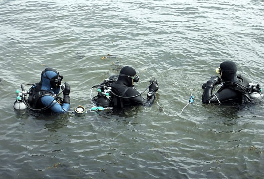 Screenshot 2026-03-15 at 230225 - N9BO℠ | Global Underwater Services Ltd Three scuba divers in full kit are floating on the surface of the water, preparing for a dive. They are wearing wetsuits and masks, and are connected by a safety line while communicating with each other.