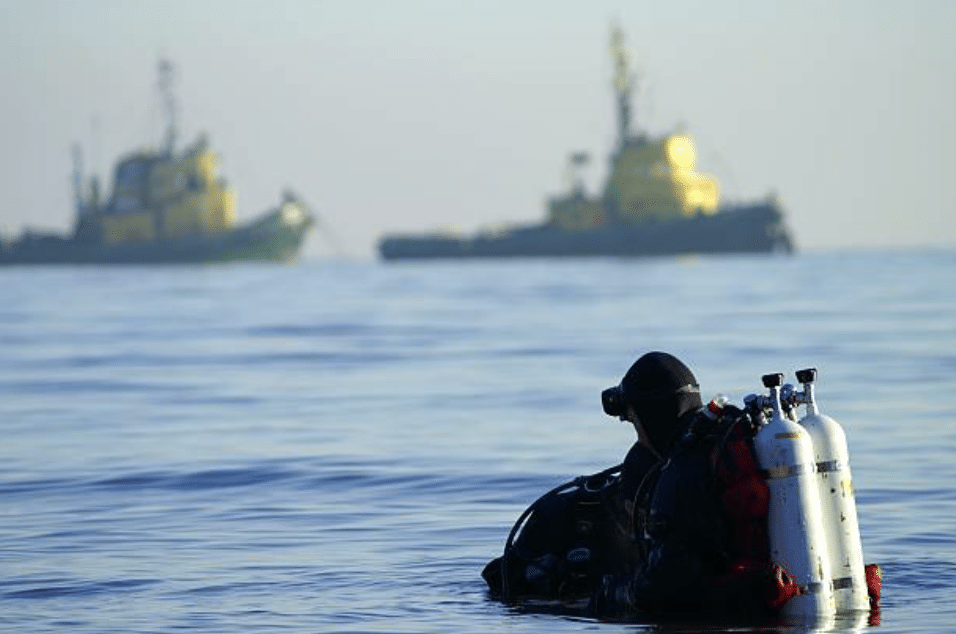 Screenshot 2026-03-15 at 230243 - N9BO℠ | Global Underwater Services Ltd A scuba diver wearing full kit with twin air tanks floats in calm water, looking towards two large ships in the distance under a clear sky.