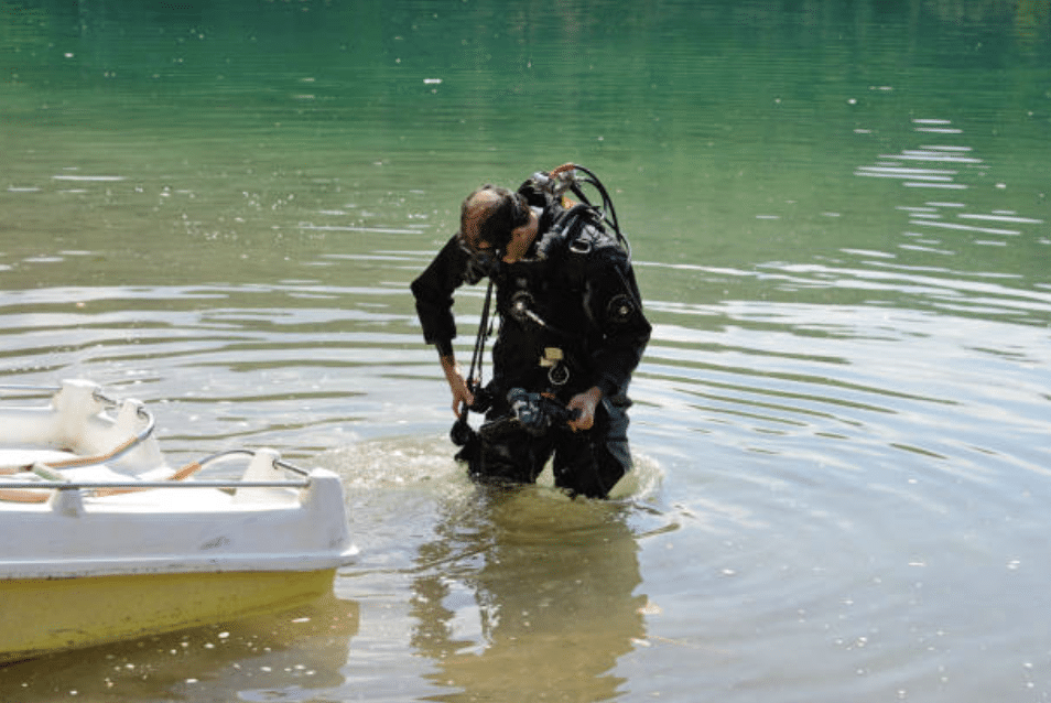 Screenshot 2026-03-15 at 230301 - N9BO℠ | Global Underwater Services Ltd A scuba diver in full kit stands knee-deep in a calm body of water near a small white boat, adjusting equipment whilst preparing for a dive.