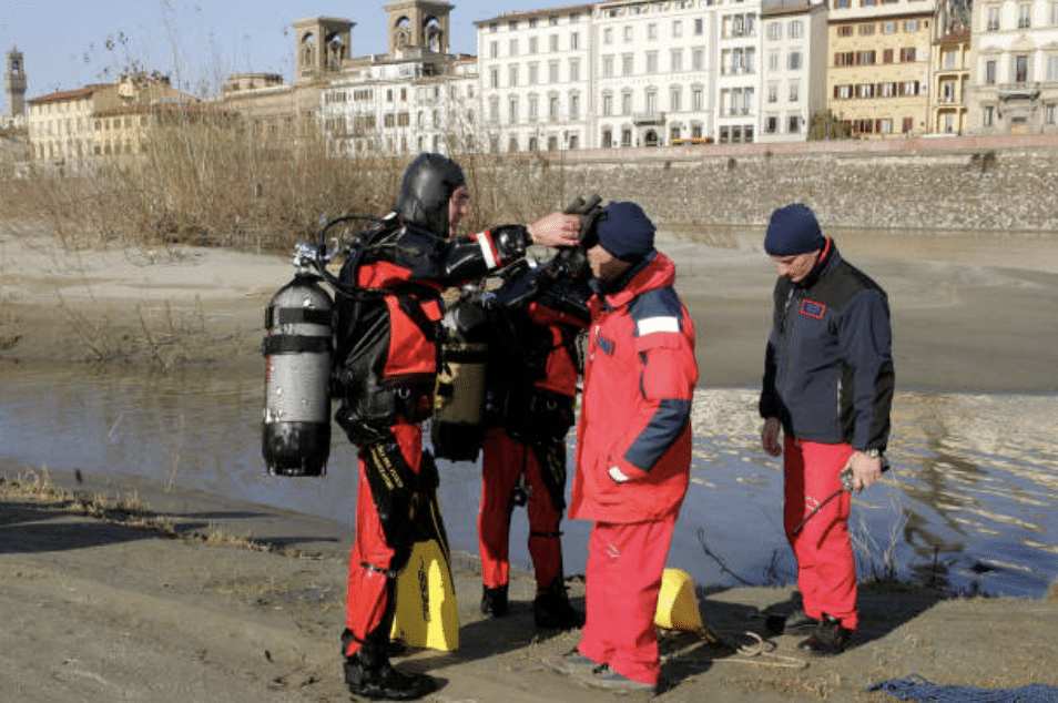 Three rescue workers in red uniforms stand near a riverbank whilst another person in a wetsuit and scuba gear adjusts equipment. Buildings and trees are visible across the river in the background.