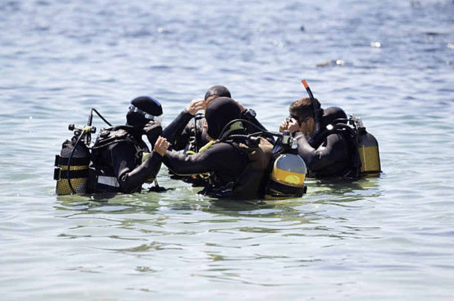 A group of scuba divers in wetsuits and diving gear gather in shallow, clear water near the shore, appearing to discuss or prepare for a dive.