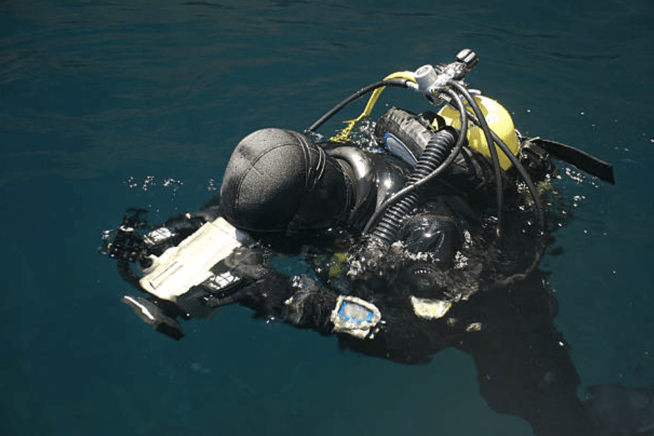 Screenshot 2026-03-15 at 233339 - N9BO℠ | Global Underwater Services Ltd A scuba diver in a black wetsuit and full diving kit is floating on the surface of dark blue water, looking down at a device or slate attached to their arm.