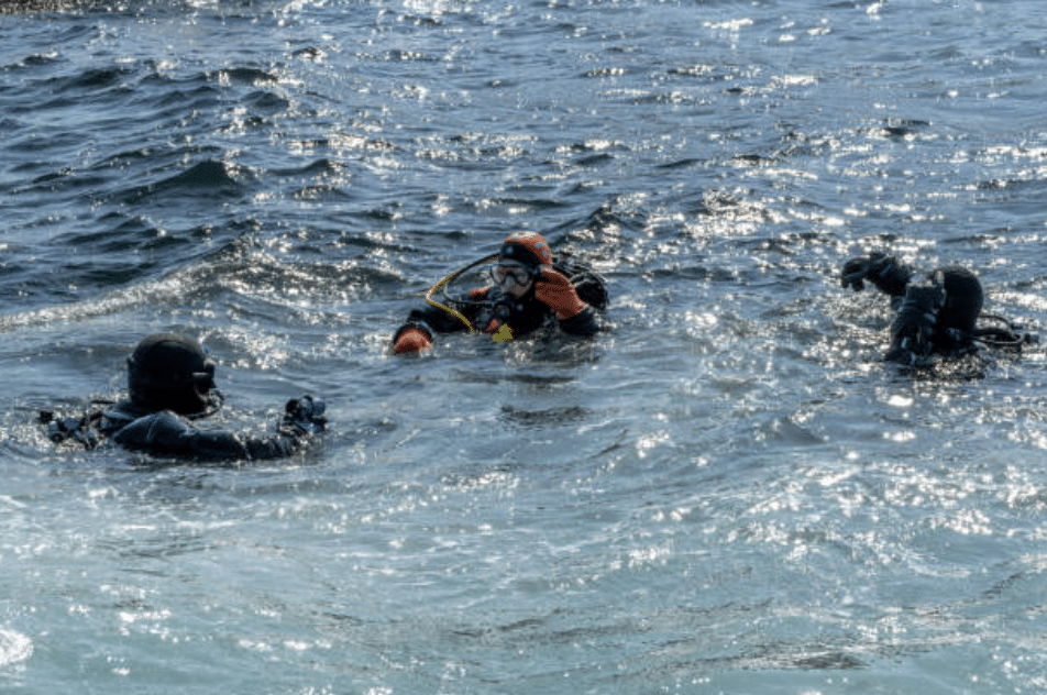 Screenshot 2026-03-15 at 233416 - N9BO℠ | Global Underwater Services Ltd Three scuba divers in wetsuits and kit float together in choppy, sunlit water, communicating with hand signals at the sea’s surface.