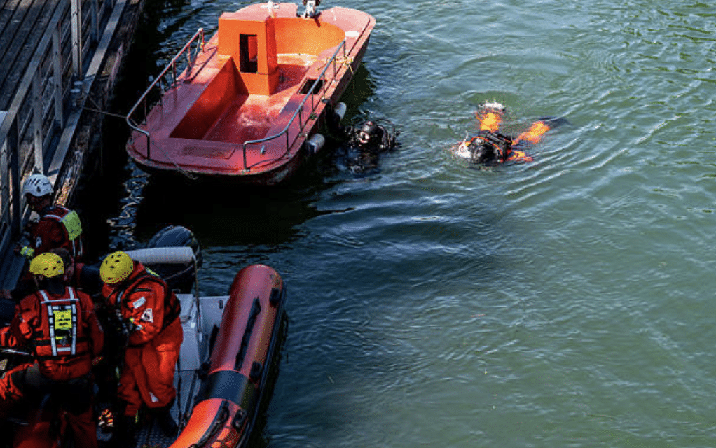 Screenshot 2026-03-15 at 234234 - N9BO℠ | Global Underwater Services Ltd Two divers in the water near a red rescue boat, with three people in orange suits and yellow helmets preparing equipment on another boat by a quay.