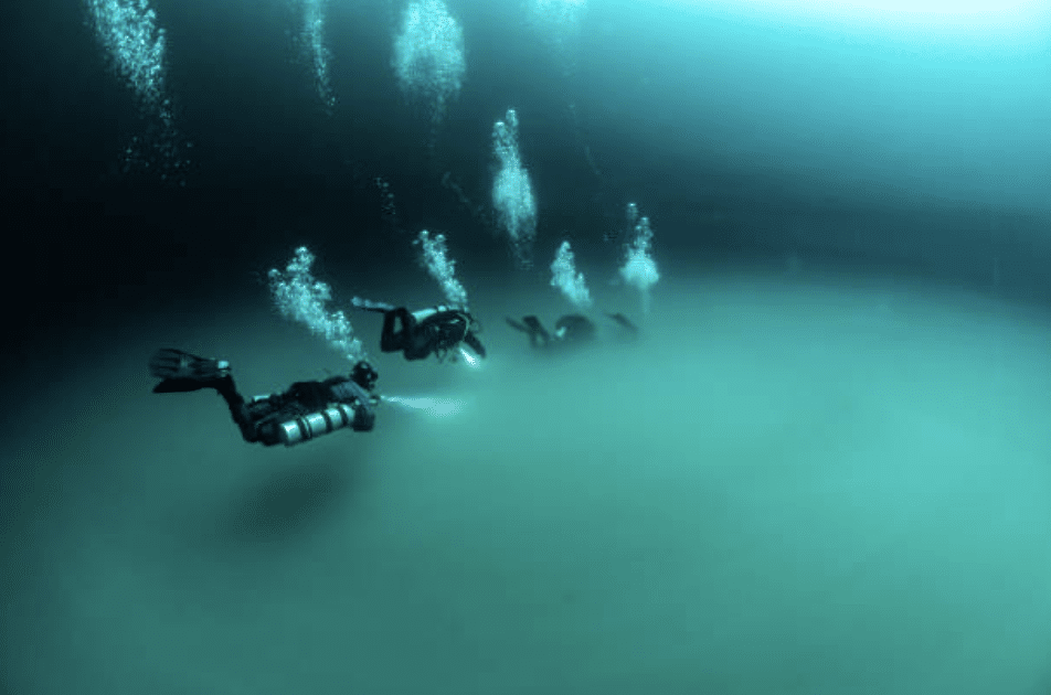 Screenshot 2026-03-15 at 235031 - N9BO℠ | Global Underwater Services Ltd A group of scuba divers swims underwater in deep, blue-green water, surrounded by bubbles and illuminated by a diver’s torch, creating a serene, otherworldly scene.