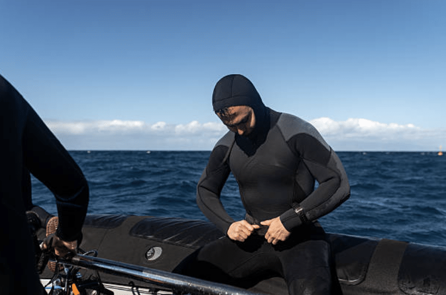 Screenshot 2026-03-16 at 000147 - N9BO℠ | Global Underwater Services Ltd A person in a black wetsuit sits on the edge of an inflatable boat, adjusting the front of their wetsuit, with the sea and a clear blue sky in the background.