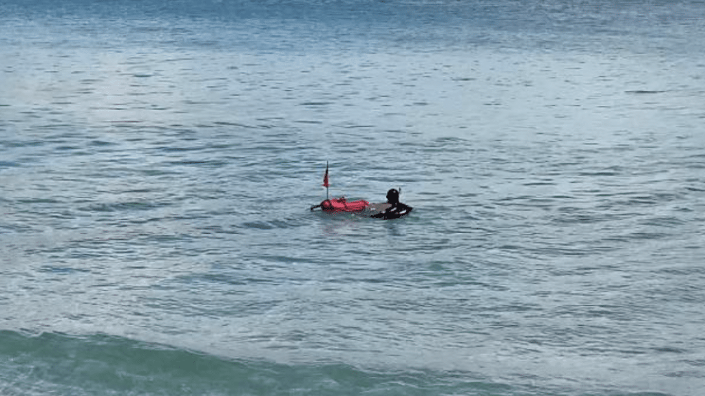 Screenshot 2026-03-16 at 000158 - N9BO℠ | Global Underwater Services Ltd A person wearing a wetsuit floats on the sea next to a small red dinghy with a flag, surrounded by calm blue water.