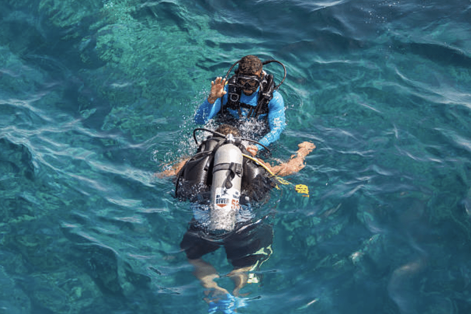 Screenshot 2026-03-16 at 001243 - N9BO℠ | Global Underwater Services Ltd Two scuba divers are in clear blue water; one appears to be instructing or assisting the other, who is partially submerged and wearing a scuba tank and kit.