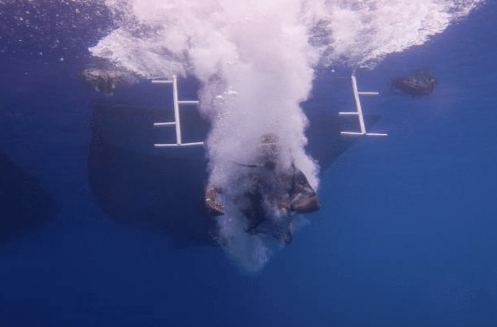 Screenshot 2026-03-16 at 002856 - N9BO℠ | Global Underwater Services Ltd A scuba diver descends feet-first into the sea, creating a trail of bubbles and splash, while other divers and the underside of a boat are visible in the background.