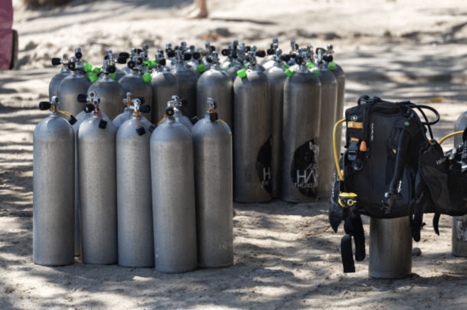 Screenshot 2026-03-16 at 004512 - N9BO℠ | Global Underwater Services Ltd Several scuba diving air cylinders and a diving gear set are grouped on sandy ground outdoors, with sunlight and partial shadows cast across the equipment.