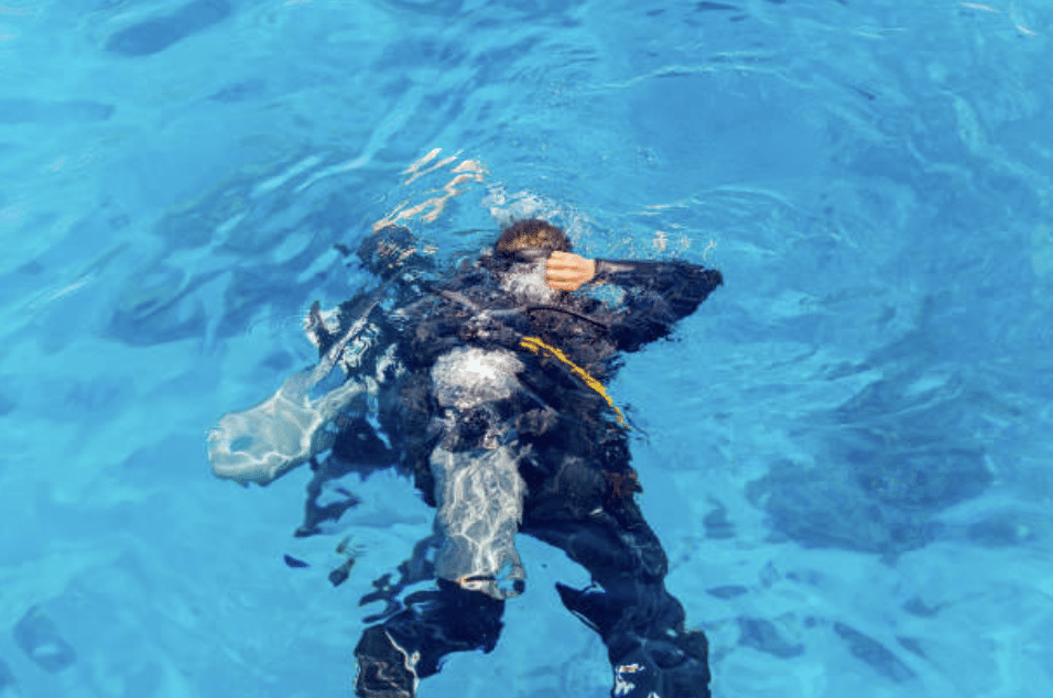 Screenshot 2026-03-16 at 004603 - N9BO℠ | Global Underwater Services Ltd A scuba diver wearing a wetsuit and kit floats face-down on the surface of clear blue water, with bubbles rising around them.