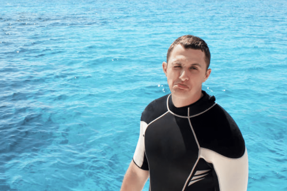 Screenshot 2026-03-16 at 004645 - N9BO℠ | Global Underwater Services Ltd A man in a black wetsuit stands in front of bright blue sea water, making a pouty face. The sun is shining, and the water appears clear and inviting.