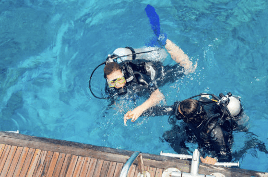 Screenshot 2026-03-16 at 010745 - N9BO℠ | Global Underwater Services Ltd Two scuba divers in wetsuits and masks float in clear blue water near a wooden jetty, with diving gear and air cylinders visible as they prepare to submerge.