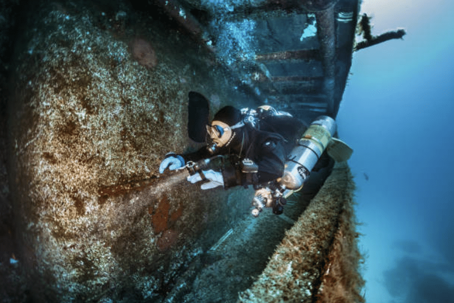 A scuba diver in full kit explores the side of a sunken shipwreck underwater, using a torch to illuminate the structure whilst surrounded by clear blue water.