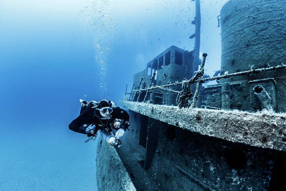 A scuba diver swims alongside a sunken, algae-covered shipwreck on the seabed, surrounded by clear blue water and rising bubbles.