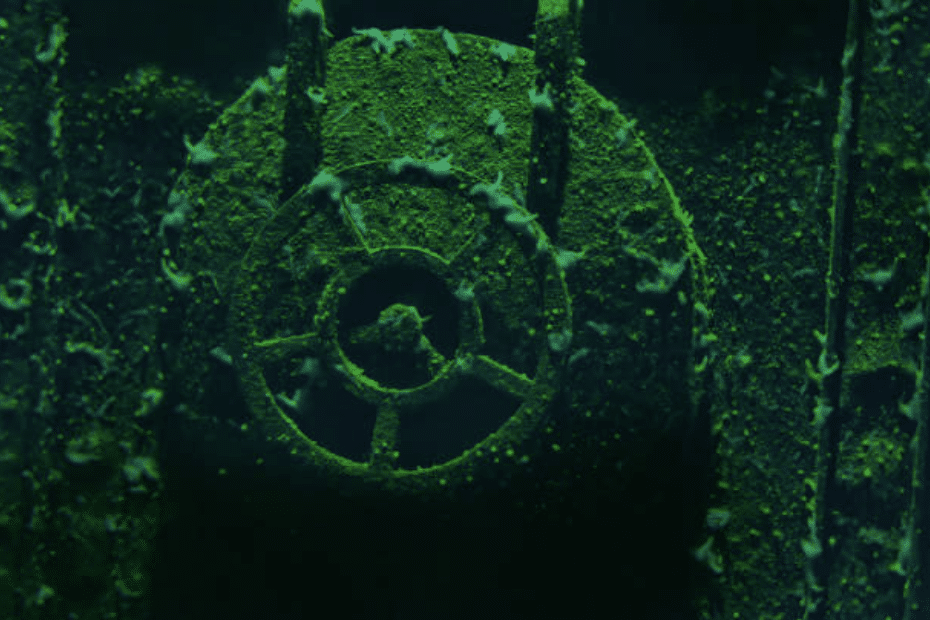 A close-up underwater view of a corroded, algae-covered valve wheel on a sunken shipwreck, illuminated by green light.