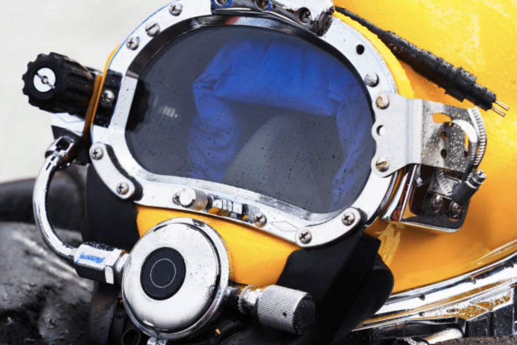 Close-up of a yellow commercial diver’s helmet with a large, reflective visor, metal fittings, and attached breathing apparatus, showing water droplets on the surface.