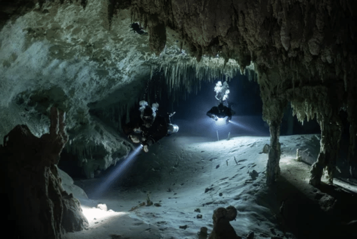Screenshot 2026-03-16 at 021946 - N9BO℠ | Global Underwater Services Ltd Two scuba divers with torches explore an underwater cave filled with stalactites and rock formations. Their beams illuminate parts of the sandy cave floor and the jagged ceiling above.