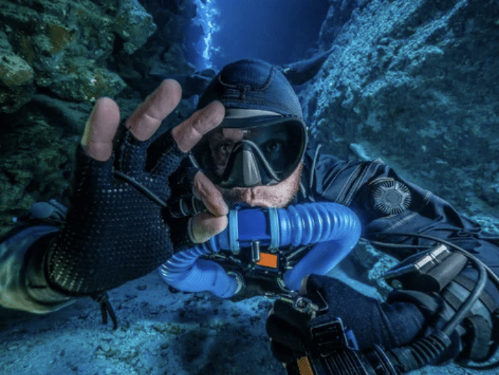 Screenshot 2026-03-16 at 022827 - N9BO℠ | Global Underwater Services Ltd A scuba diver wearing a full wetsuit, gloves, and a mask makes an OK hand signal underwater between rocky formations, illuminated by blue light from above.