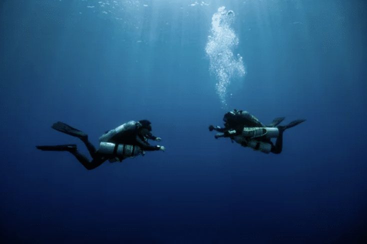Screenshot 2026-03-16 at 022843 - N9BO℠ | Global Underwater Services Ltd Two scuba divers in full kit face each other underwater, surrounded by deep blue sea with sunlight streaming down from above. Bubbles rise from one diver as they interact.