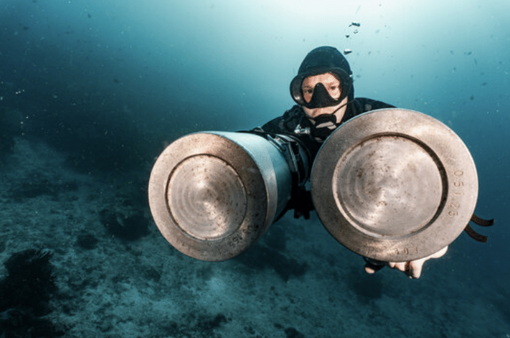 Screenshot 2026-03-16 at 023816 - N9BO℠ | Global Underwater Services Ltd A scuba diver underwater holds two large metal cylinders, possibly scuba tanks or propulsion devices, while wearing a mask and wetsuit. The seabed and blue water are visible in the background.