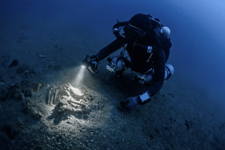 Screenshot 2026-03-16 at 024145 - N9BO℠ | Global Underwater Services Ltd A scuba diver in dark kit uses a torch to examine bones or artefacts on the seabed in deep, blue water.