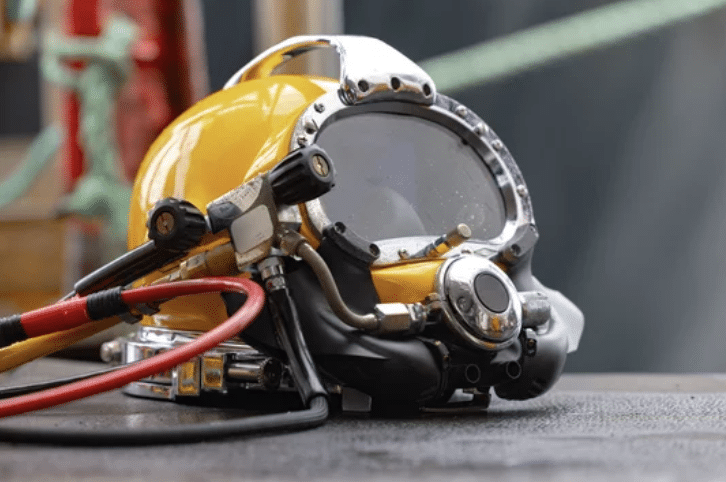 A yellow commercial diver’s helmet with a clear faceplate, metal fittings, and attached hoses rests on a textured surface, with blurred industrial equipment visible in the background.