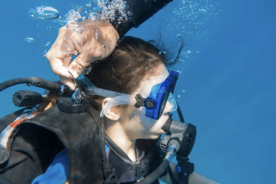 Screenshot 2026-03-16 at 191015 - N9BO℠ | Global Underwater Services Ltd A person underwater wearing a scuba mask and breathing apparatus is being assisted by another hand adjusting their mask strap. Bubbles rise around them in the clear blue water.