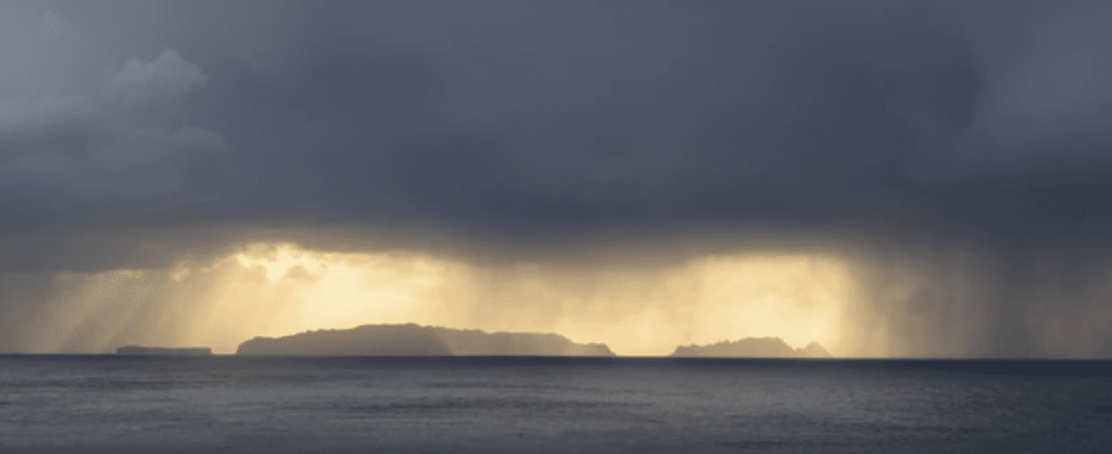 Screenshot 2026-03-16 at 201131 - N9BO℠ | Global Underwater Services Ltd Dark storm clouds loom over distant islands as sunlight breaks through, creating dramatic beams of light over the ocean and illuminating the rain in the background.