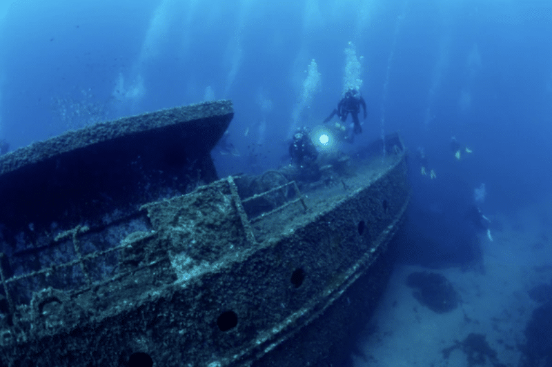 Scuba divers explore a large, submerged shipwreck on the seabed, with bubbles rising to the surface and marine life visible around the rusted, algae-covered structure.