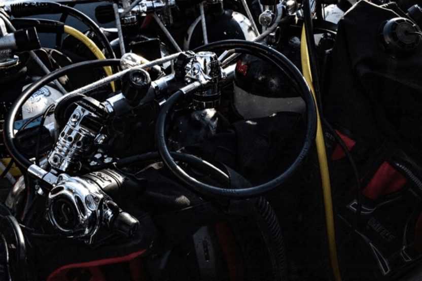Screenshot 2026-03-16 at 210941 - N9BO℠ | Global Underwater Services Ltd A close-up of scuba diving equipment, including regulators, hoses, and cylinders, tangled together in a pile with some yellow and black hoses visible among the kit.