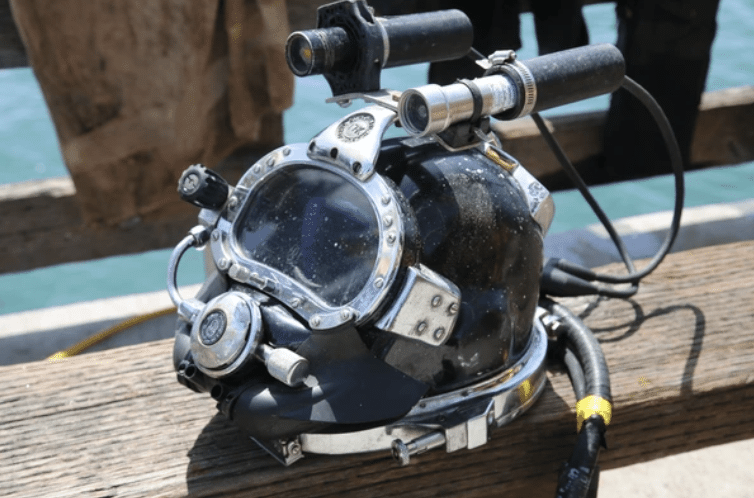 Screenshot 2026-03-16 at 211001 - N9BO℠ | Global Underwater Services Ltd A close-up of a black and silver commercial diving helmet with attached lights and hoses, resting on a wooden surface near water on a sunny day.