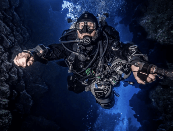 Screenshot 2026-03-16 at 211631 - N9BO℠ | Global Underwater Services Ltd A scuba diver in full kit swims underwater between rocky walls, surrounded by bubbles and illuminated by blue light filtering from above.