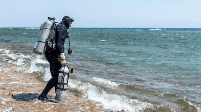 Screenshot 2026-03-16 at 213628 - N9BO℠ | Global Underwater Services Ltd A scuba diver in a wetsuit and mask stands at the edge of the sea, holding two oxygen cylinders, preparing to enter the water on a sunny day. Waves lap at the shore in the background.