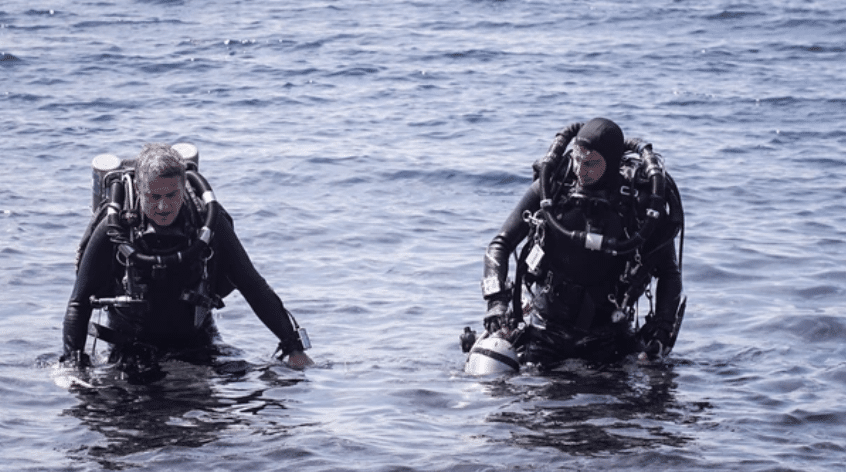 Screenshot 2026-03-16 at 213645 - N9BO℠ | Global Underwater Services Ltd Two scuba divers in full kit stand waist-deep in the sea, facing forward, with blue water and gentle waves surrounding them.