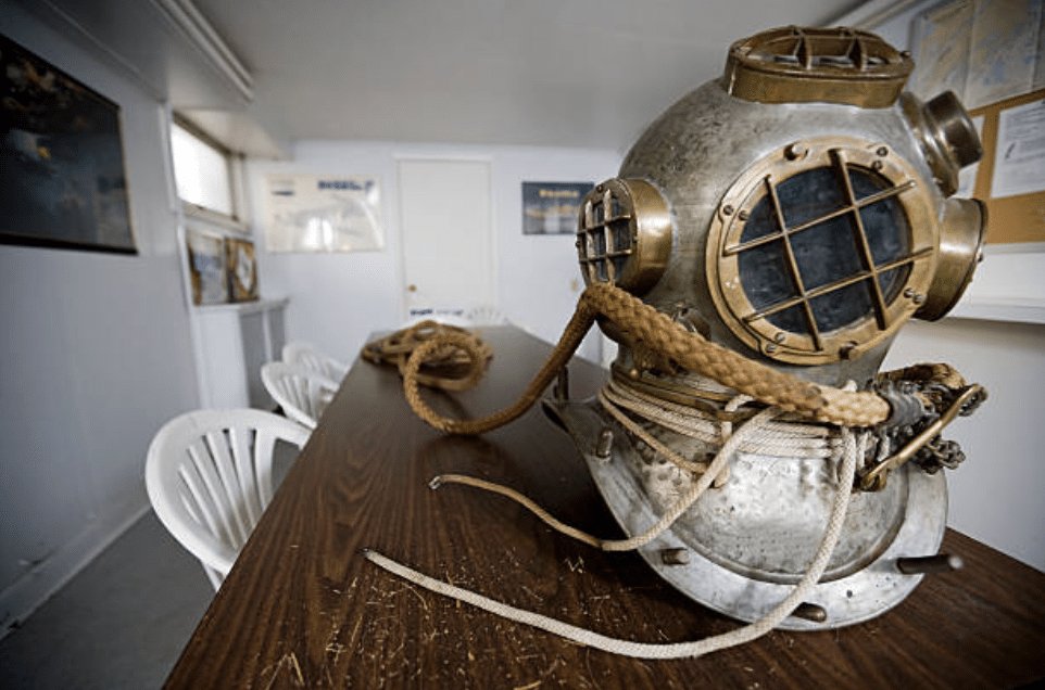 Screenshot 2026-03-16 at 214453 - N9BO℠ | Global Underwater Services Ltd A vintage metal diving helmet with thick ropes rests on a long wooden table in a room with white chairs and nautical posters on the walls.