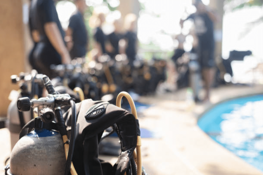 Screenshot 2026-03-16 at 214523 - N9BO℠ | Global Underwater Services Ltd A row of scuba diving cylinders and equipment is lined up beside a swimming pool, with several people standing and preparing in the blurred background.